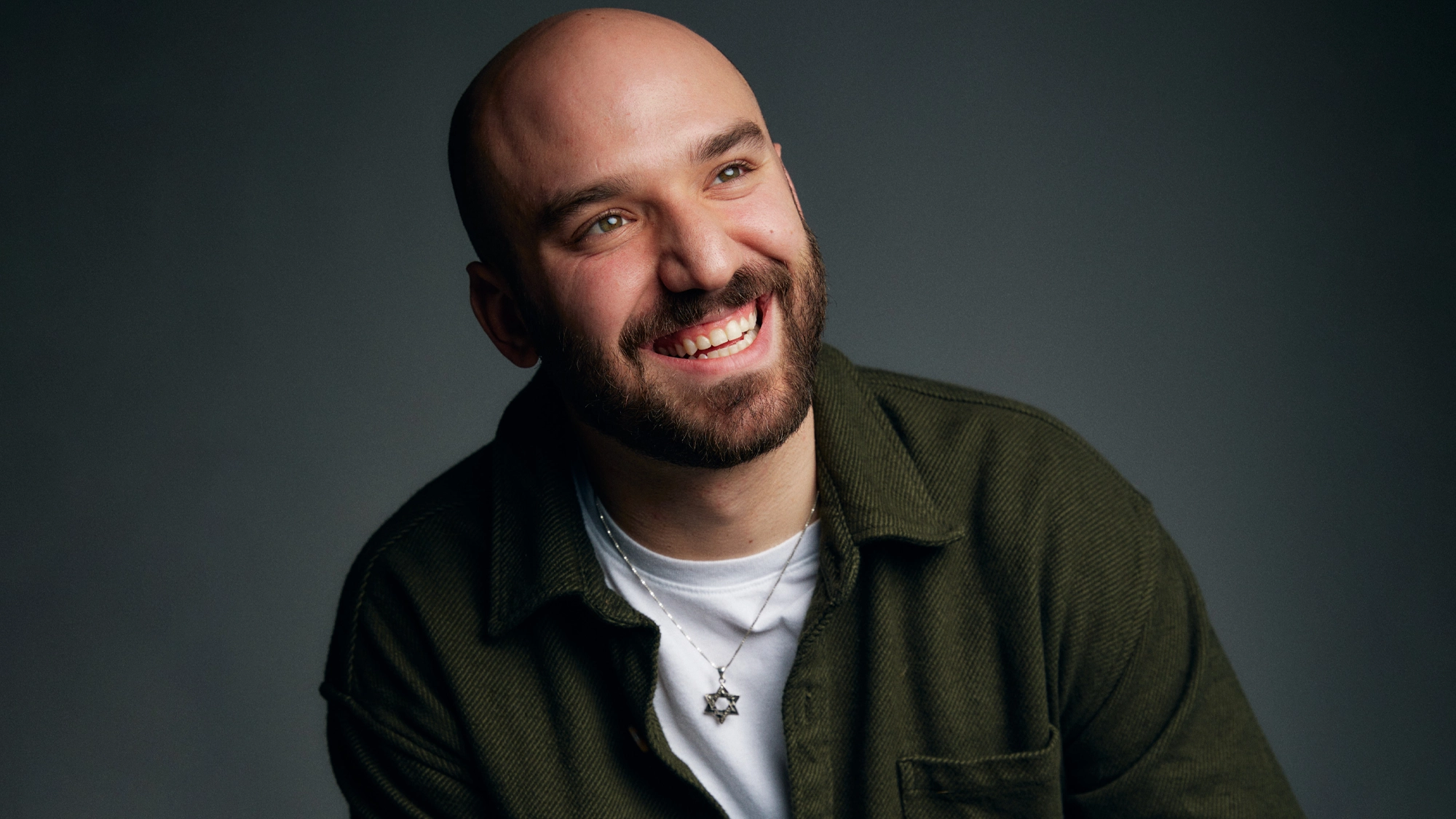 A smiling bald man with a beard, Ari Axelrod, wears a dark green jacket over a white shirt and a Star of David necklace. He looks up and to the right against a dark background, radiating the spirit of L’chaim—To Life.