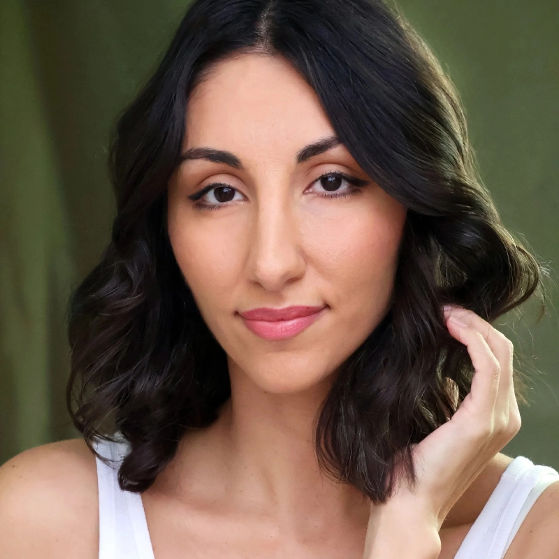 Kristina Asriyan, with medium skin tone and wavy dark brown hair, gazes gently at the camera. She wears a white tank top, lightly touching her hair with her right hand against a softly blurred green background.