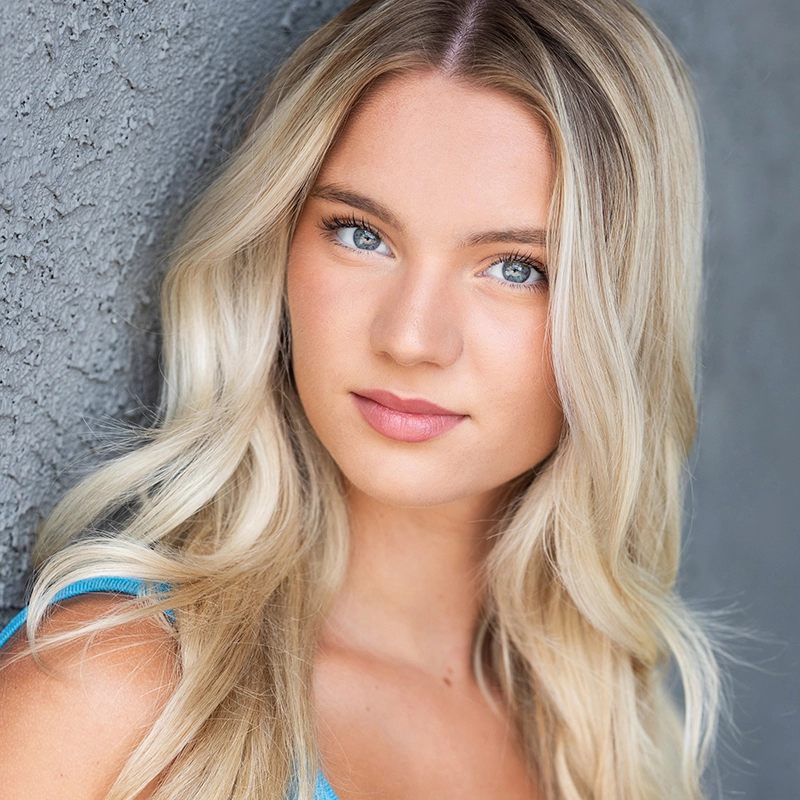 Audrey Graves, a young woman with long, wavy blonde hair and blue eyes, stands against a textured gray wall. She wears a sleeveless blue top and looks directly at the camera with a calm, neutral expression and natural makeup.
