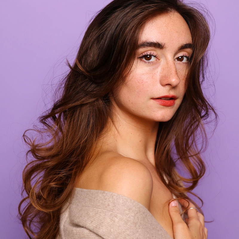 Madison August, with long, wavy brown hair, stands against a purple background. She gazes confidently at the camera in a beige off-shoulder top, her natural makeup highlighting her shoulder and collarbone.