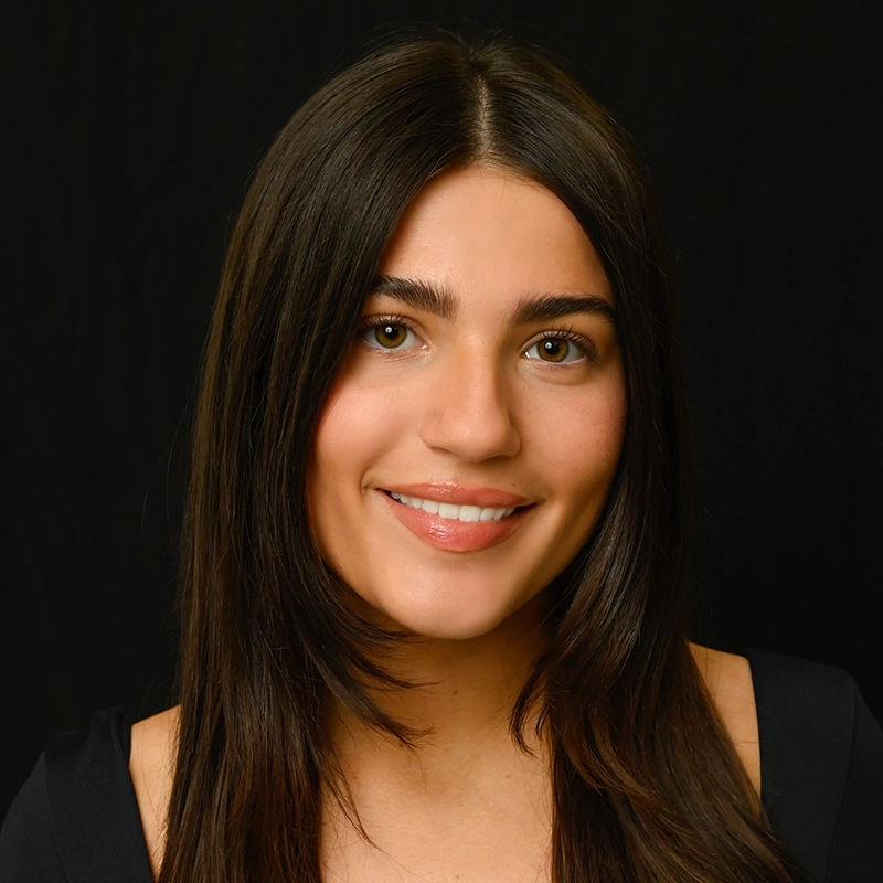 A young woman with straight, long dark brown hair and hazel eyes smiles softly. She wears a black top and is posed against a plain black background, looking directly at the camera. The lighting is bright and evenly highlights her face.