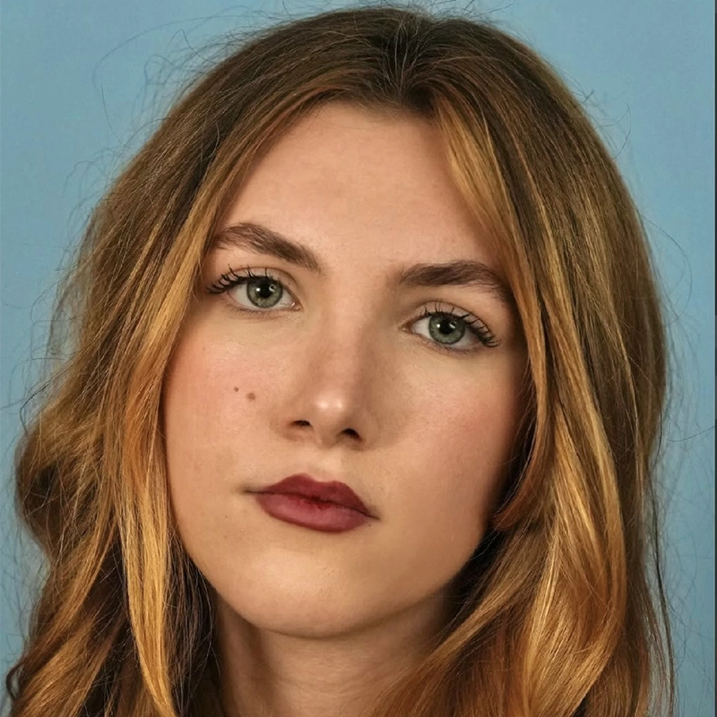 A young woman with light skin and long, wavy, light brown hair stares directly at the camera with a neutral expression. She has green eyes, bold eyebrows, and wears natural makeup with dark rose lipstick. Farida Wahby stands against a plain light blue background.