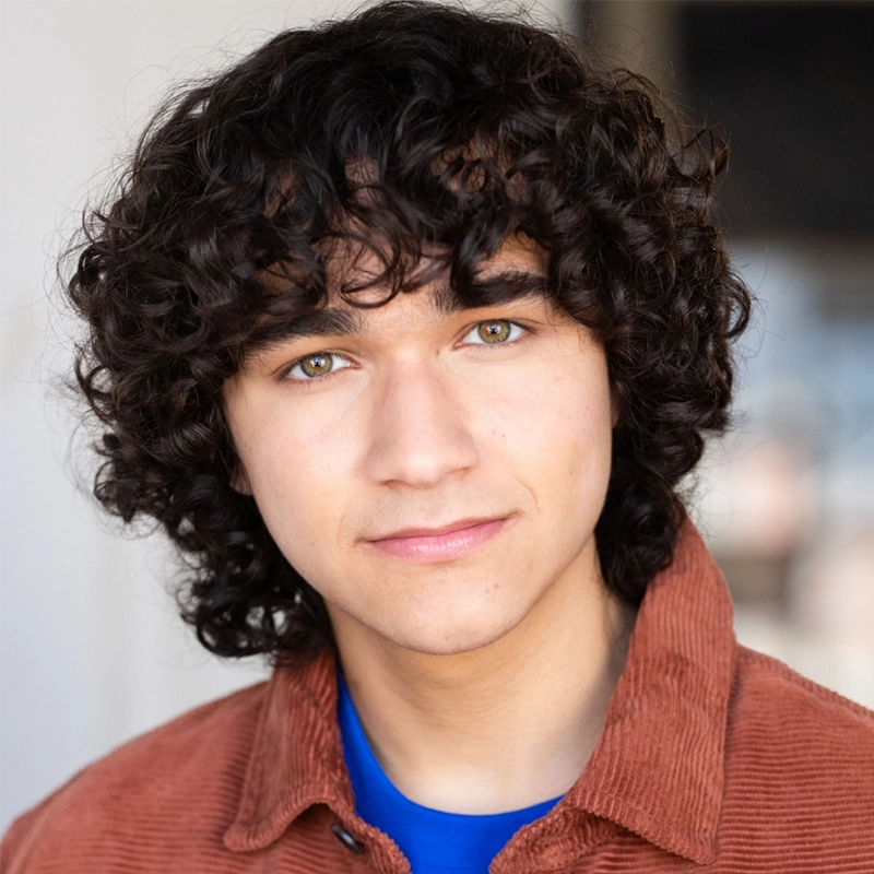 A young person with medium-length curly dark hair and hazel eyes looks into the camera, smiling slightly. They wear a rust-colored corduroy jacket over a blue shirt. The background is softly blurred in neutral tones.