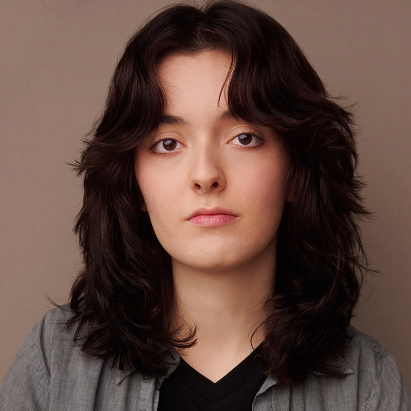 Danté X Johnson, with shoulder-length dark wavy hair and fair skin, looks directly at the camera with a neutral expression. They are wearing a grey button-up shirt over a black top against a plain beige background.