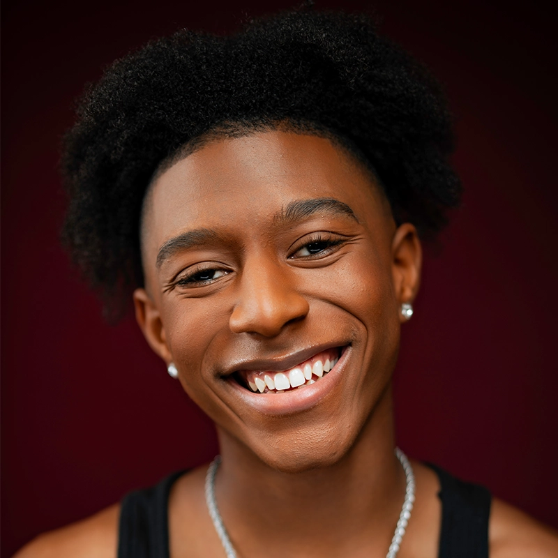 A young person with medium brown skin, short curly hair, and a bright smile poses against a dark red background. Danté X Johnson wears a black tank top, a silver necklace, and small stud earrings, exuding warmth and confidence.
