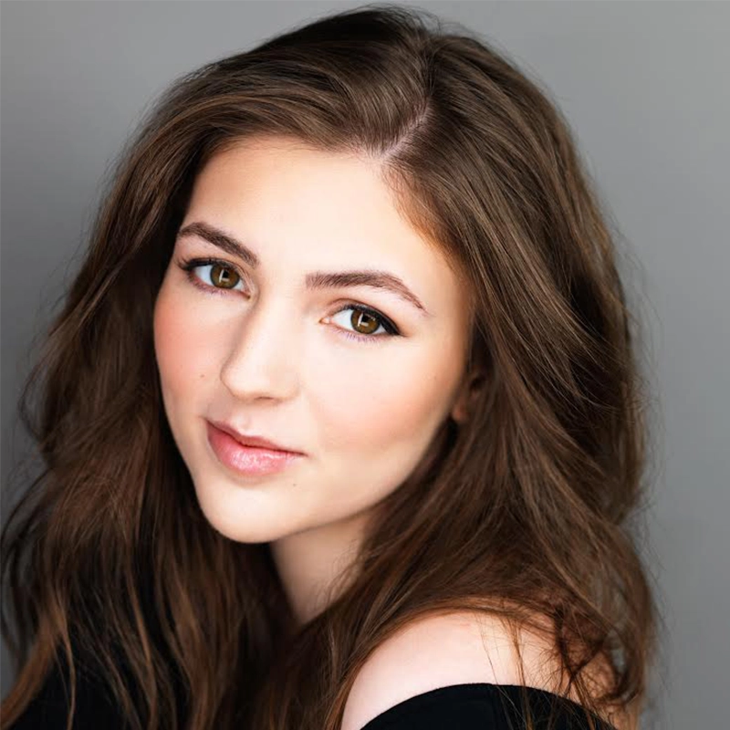 A young woman with long, wavy brown hair and fair skin gazes softly at the camera. She has brown eyes, defined eyebrows, and subtle makeup. Her lips are lightly glossed, and she wears an off-the-shoulder black top against a gray background.