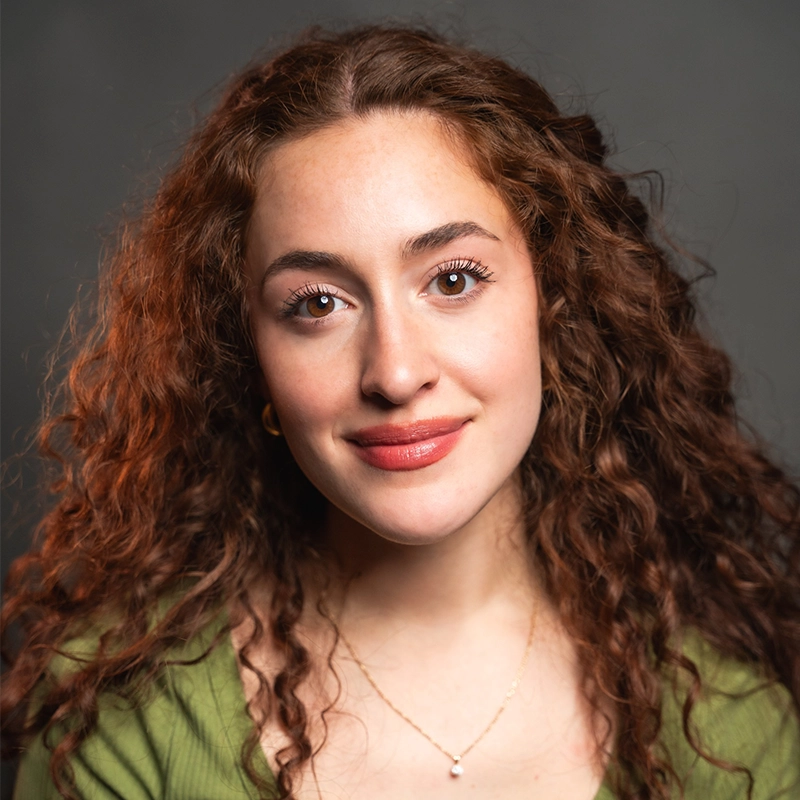 A young woman with long, curly auburn hair poses against a dark background. She has fair skin, brown eyes, and wears natural makeup with pink lipstick. She is dressed in a green top and a delicate gold necklace with a small pendant.