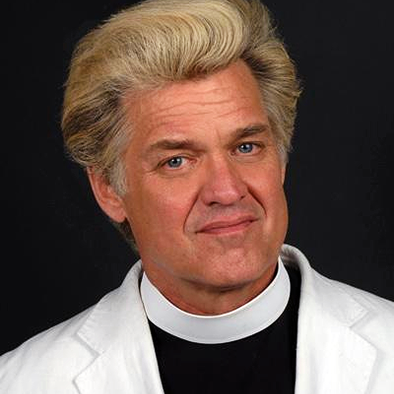A man with thick, swept-back blond hair wearing a white clergy collar and a white jacket over a black shirt, Reverend Billy looks at the camera with a slight smile against a dark background.