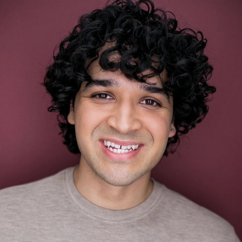A young person with curly dark hair and medium skin tone smiles warmly at the camera. Jorge Blakely wears a light beige shirt and stands against a solid maroon background. The lighting is bright and even, highlighting his friendly expression.