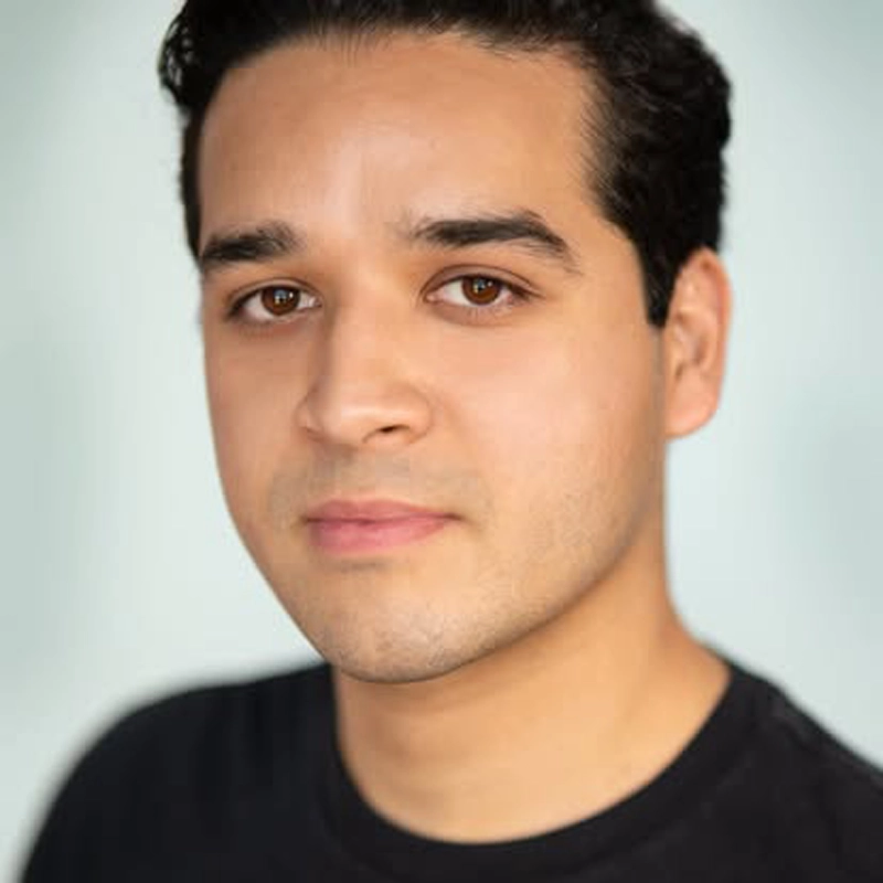Jorge Blakely, a young man with short, dark hair and brown eyes, looks directly at the camera with a neutral expression. He is wearing a black crew-neck shirt. The pale blue, softly blurred background draws focus to his face.