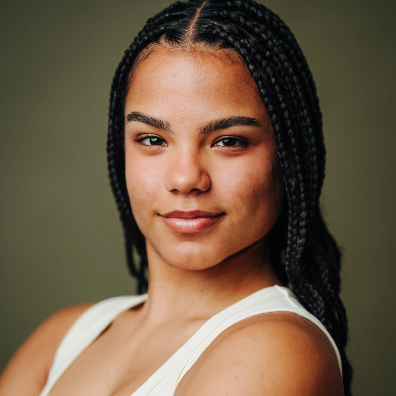 A young woman with medium skin tone and neatly braided hair looks confidently at the camera. She wears a sleeveless white top and has a calm, slight smile. The background is a plain, muted olive green.
