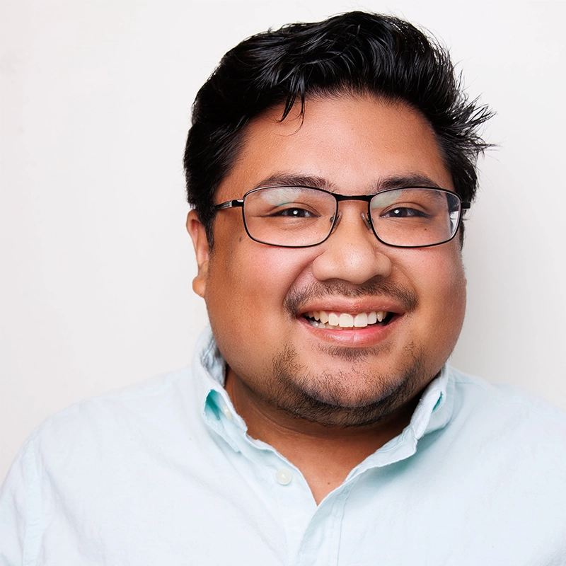 A smiling man with short black hair, a trimmed beard, and glasses wears a light blue button-up shirt. He is facing forward against a plain white background, appearing cheerful and approachable, reminiscent of Gabriella Santalla’s positive energy.
