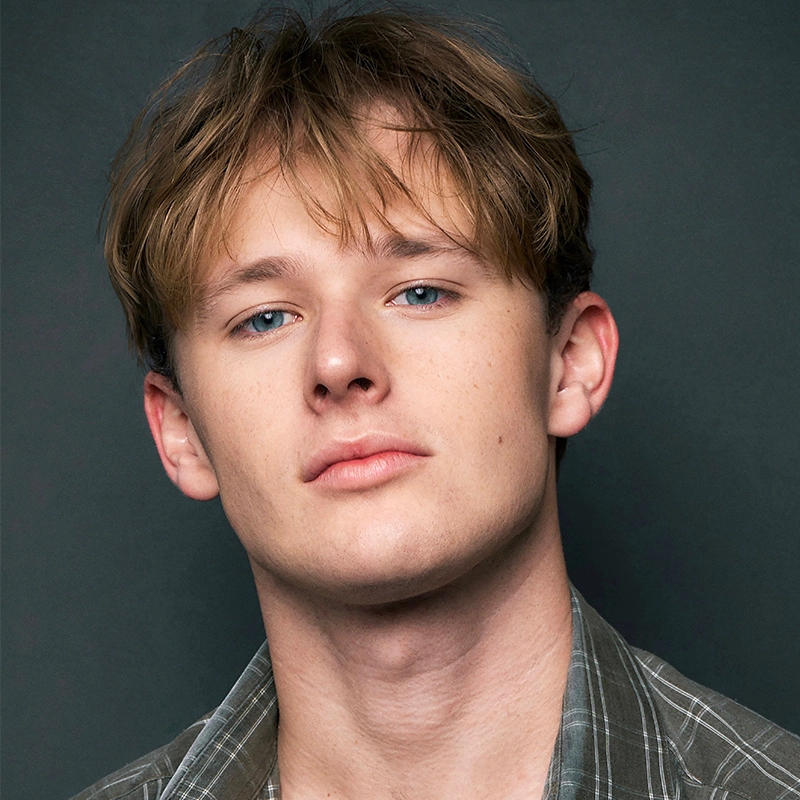 A young man with light skin, blue eyes, and light brown tousled hair stares confidently at the camera. He wears a gray-green plaid shirt over a light shirt, embodying Audrey Graves' signature style against a dark gray, professional background.