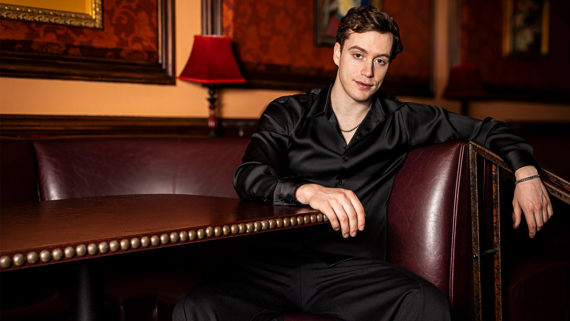 A young man with short brown hair, wearing a black satin shirt and black pants, sits comfortably in a maroon leather booth in a warmly lit room with red patterned wallpaper and decorative lamps, looking at the camera with a relaxed expression.