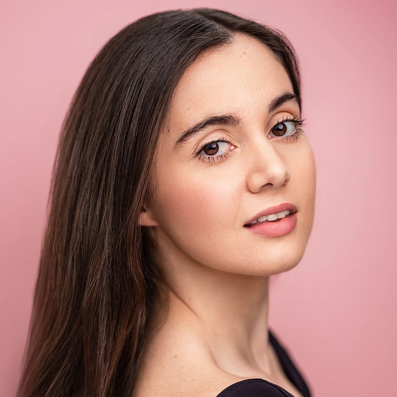 Brooke Cassatto, a young woman with long straight brown hair and natural makeup, poses against a soft pink background. She wears a black top, looking gently over her shoulder with smooth skin and a confident expression.