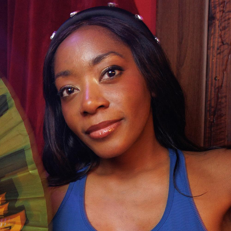 Nonye Brown-West, a woman with dark skin and long, dark hair, wears a blue sleeveless top and a black headband with pearls. She faces the camera with a slight smile, holding a green fan. Red curtains and wooden panels form the backdrop.