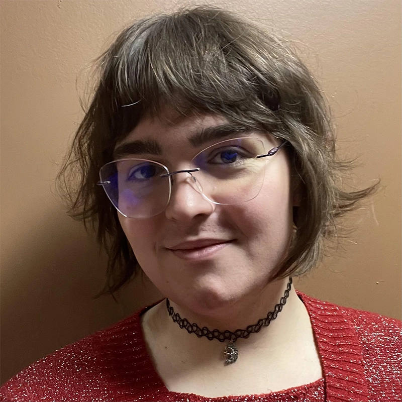 Genevieve Inez, with short, wavy brown hair and glasses, smiles slightly. She wears a black choker necklace with a crescent moon charm and a red knit sweater against a plain light brown wall.