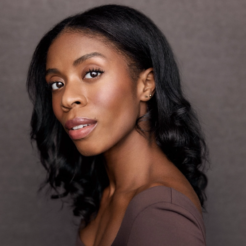 A woman with long, wavy black hair looks slightly over her shoulder. She has smooth brown skin, subtle natural makeup, and wears a fitted brown top. The background is a soft, neutral gray, giving this Runako Campbell portrait a professional feel.