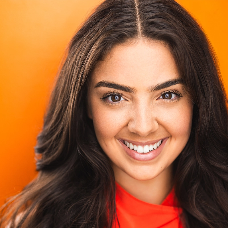 A young woman, Farida Wahby, with long dark hair smiles warmly at the camera. She has thick eyebrows, clear skin, and is wearing a bright orange top against a solid orange background, creating a vibrant, cheerful atmosphere.