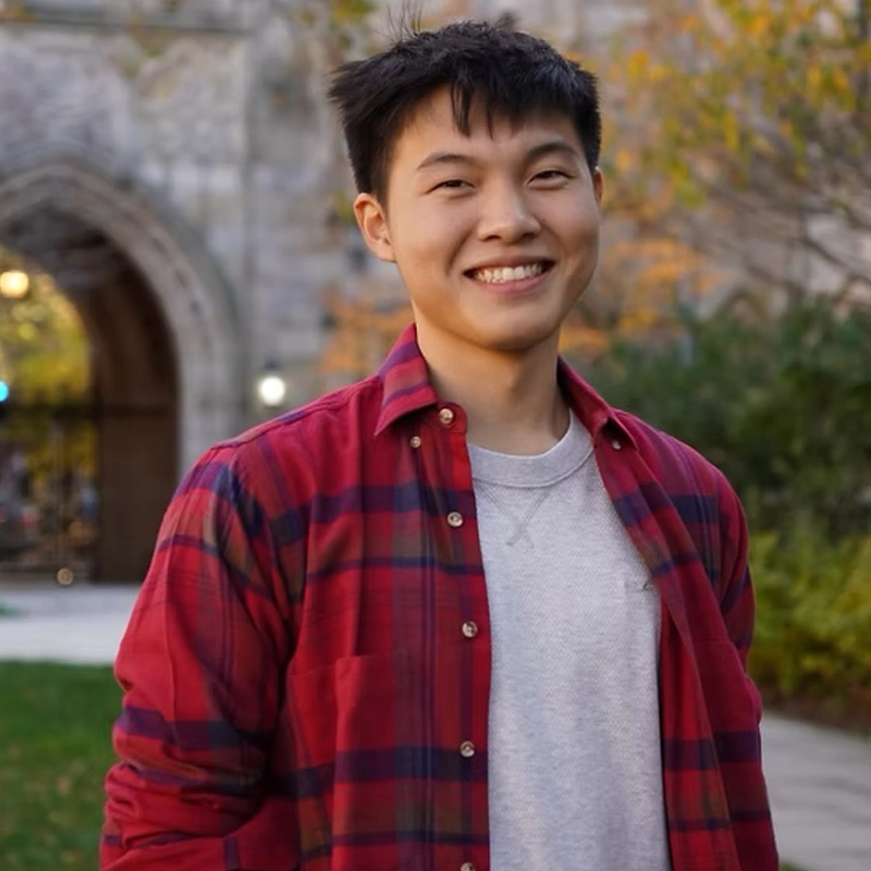 A young man with short black hair smiles outdoors. He wears a red plaid shirt over a light gray t-shirt. Behind him are green trees, grass, and a stone building with an arched entrance, suggesting a college or university campus.
