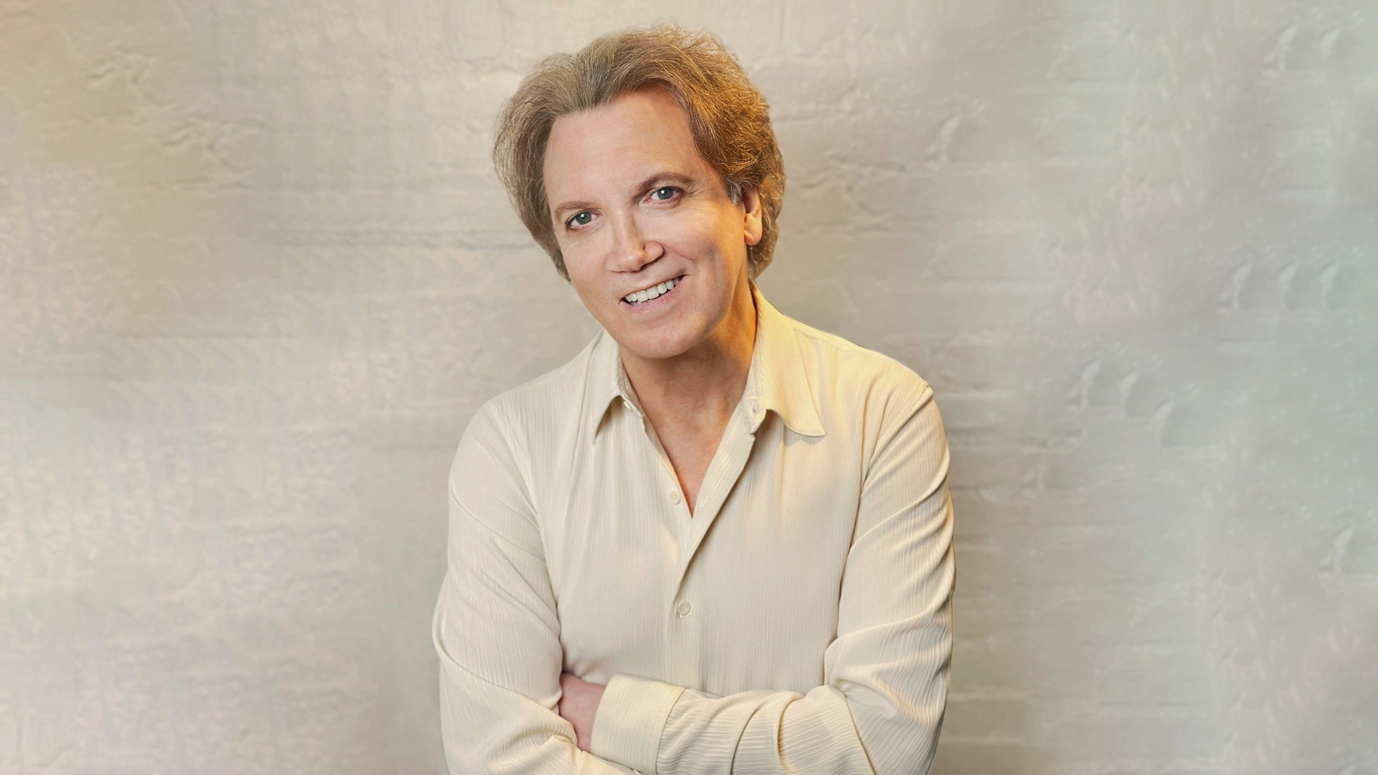 A smiling man with light brown hair stands against a textured white wall. He is wearing a cream-colored button-up shirt with rolled sleeves, his arms loosely crossed, radiating warmth—much like Johnny Mercer when he sings in a welcoming atmosphere.