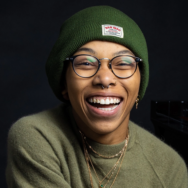 A person smiles broadly, showing teeth. They wear round glasses, a green beanie with a clover patch, and a matching green sweater. Multiple gold necklaces, earrings, and nose piercings stand out against the dark, blurred background.