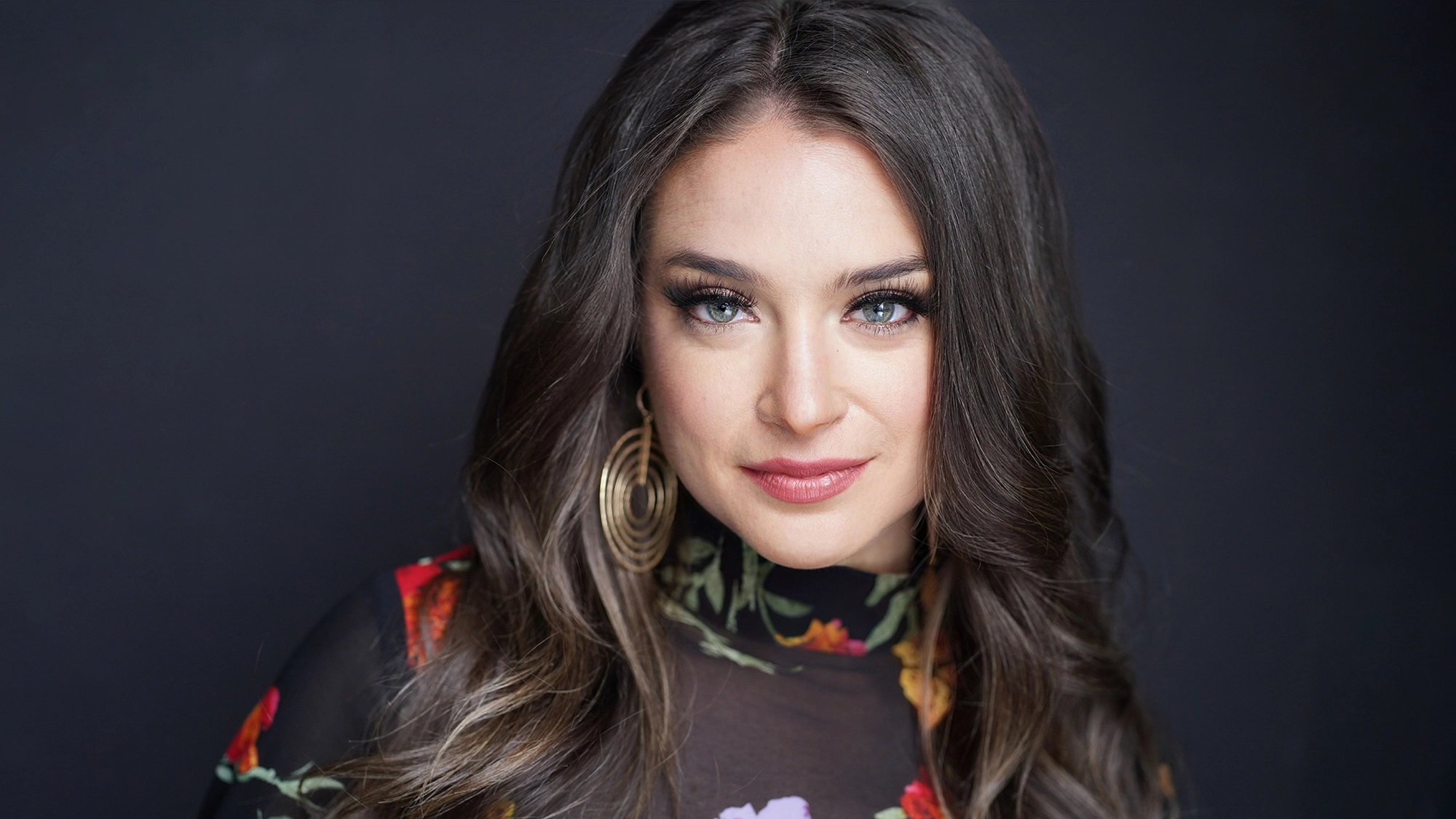 A woman with long brown hair and blue-green eyes gazes into the camera, smiling slightly. She wears large gold hoop earrings and a dark, sheer floral top—her look perfect for a late-night stroll or being caught under the mistletoe.