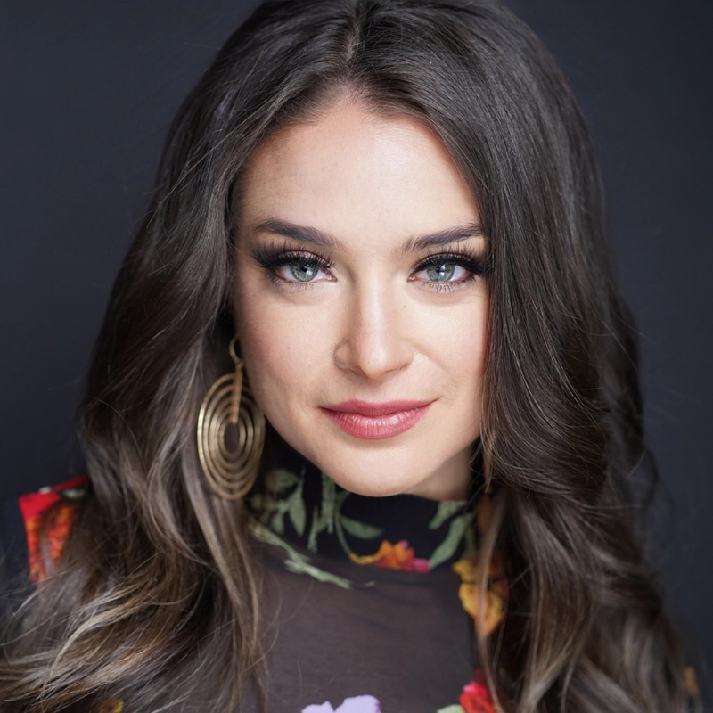 A woman with long, wavy brown hair and blue eyes gazes confidently at the camera. She wears bold makeup, large gold spiral earrings, and a sheer black top with a colorful floral pattern. The background is dark and plain.