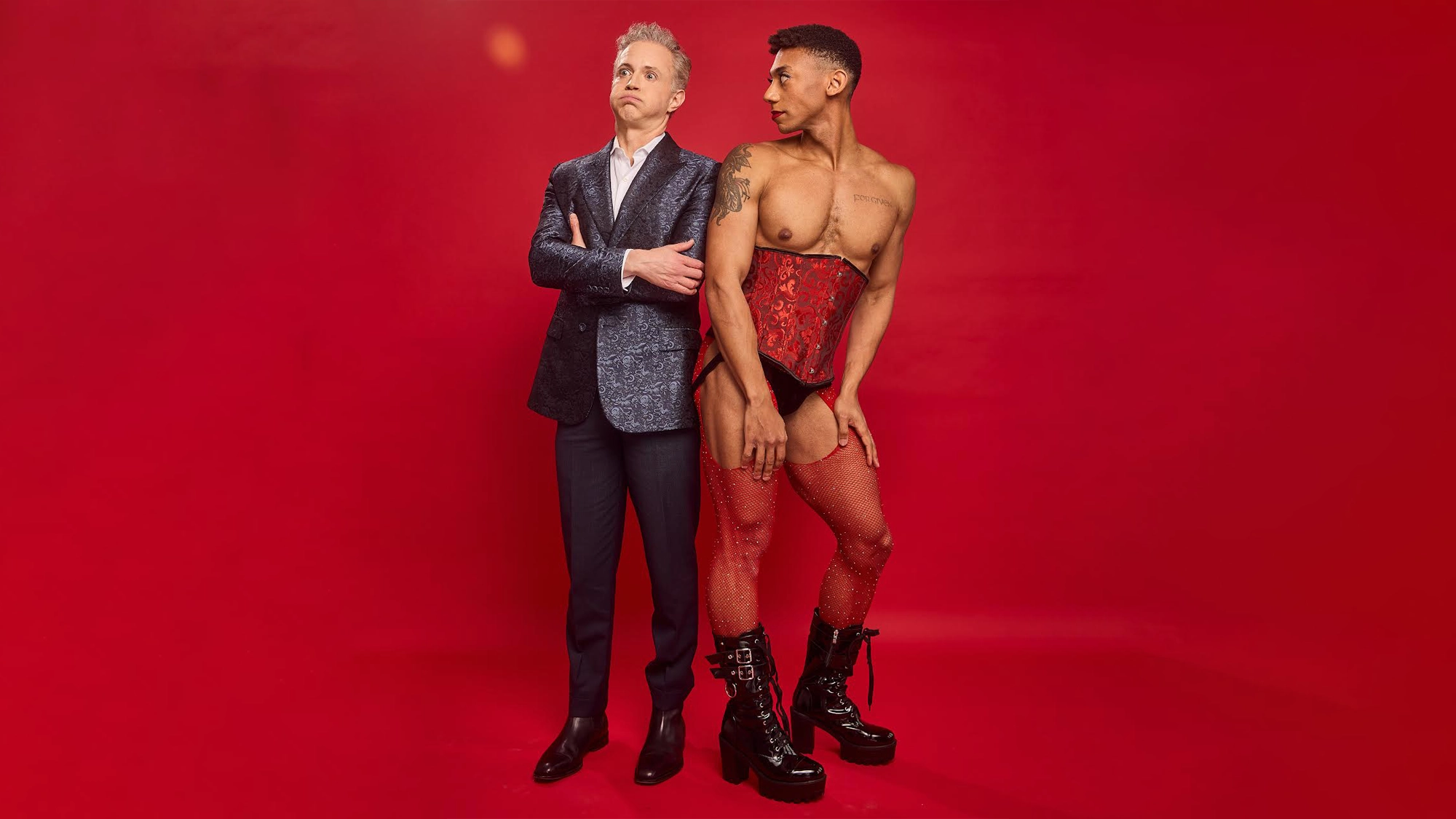 Two people stand against a solid red background. On the left, Landon Stovall in a shiny blue suit with arms crossed looks sideways. On the right, Tom Guthrie poses confidently in a red corset, fishnet stockings, and black platform boots for The Door.