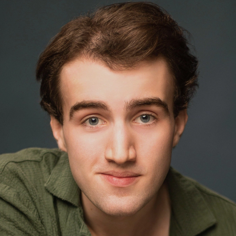 A young man with fair skin, short brown hair, and blue eyes, wearing a green collared shirt, looks directly at the camera with a neutral expression. The background is plain and dark, keeping the focus on his face.
