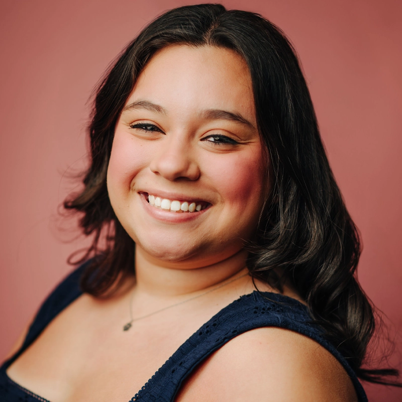 A young woman with medium skin tone and long, wavy brown hair smiles warmly at the camera. She is wearing a navy blue sleeveless top and a delicate necklace. The background is a solid, soft pink color.