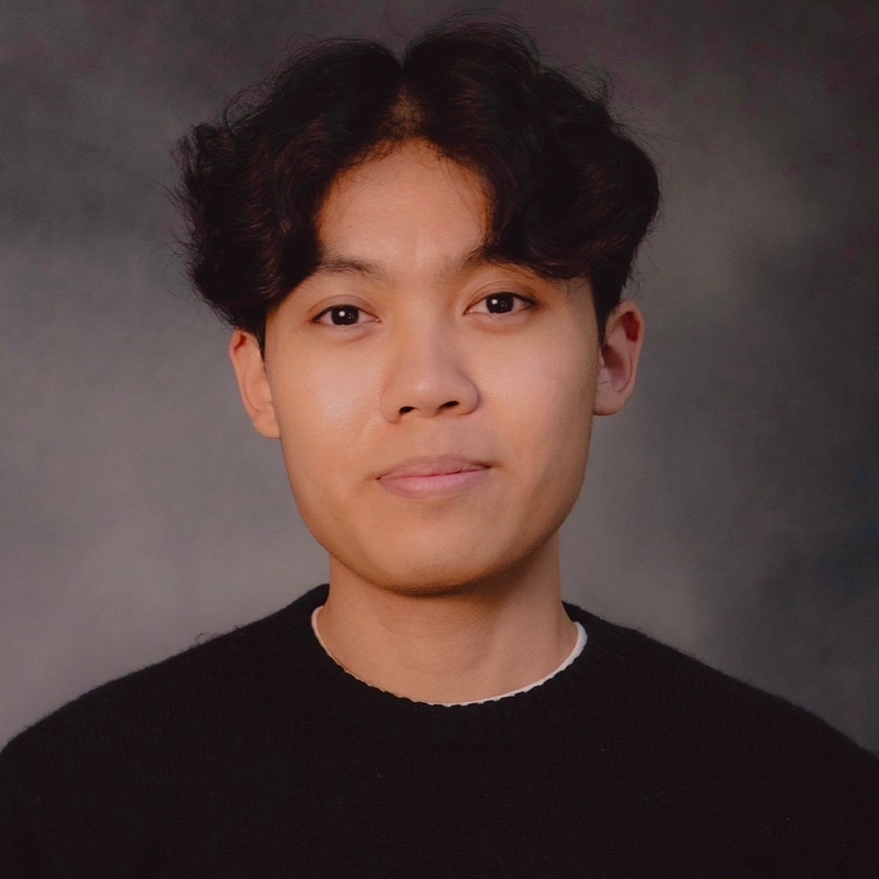 A young person with short, wavy dark hair and light brown skin looks at the camera with a neutral expression. They wear a black sweater over a white shirt. The background is a muted, gradient grey.