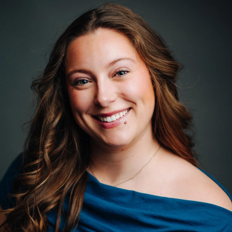 Mackenzie Cunnane, a woman with long, wavy brown hair, smiles warmly at the camera. She has blue eyes, a beauty mark above her lip, and is wearing a blue off-the-shoulder top against a dark, softly blurred background.