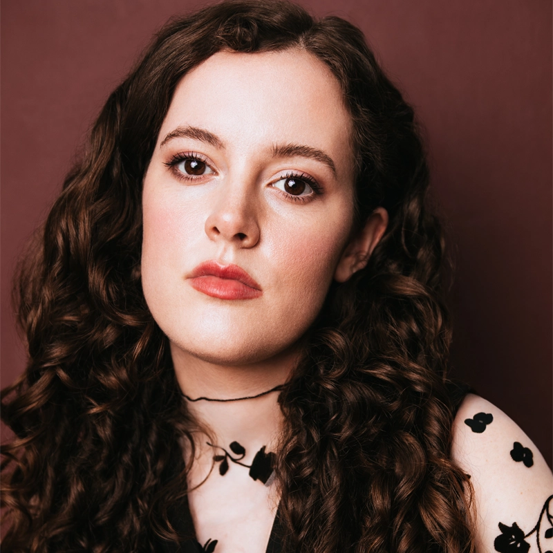 Amanda D'Amico, a young woman with long, dark brown curly hair, poses calmly against a maroon background. She has fair skin, soft makeup, and wears a black top with a floral pattern and a thin black choker. Her expression is neutral and composed.
