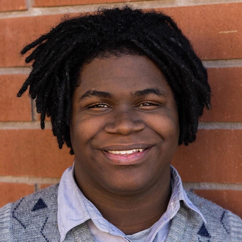 Danté X Johnson, a young person with dark skin and short, twisted dreadlocks, smiles warmly at the camera. They wear a light blue collared shirt under a gray sweater, set against a wall of reddish-brown bricks.