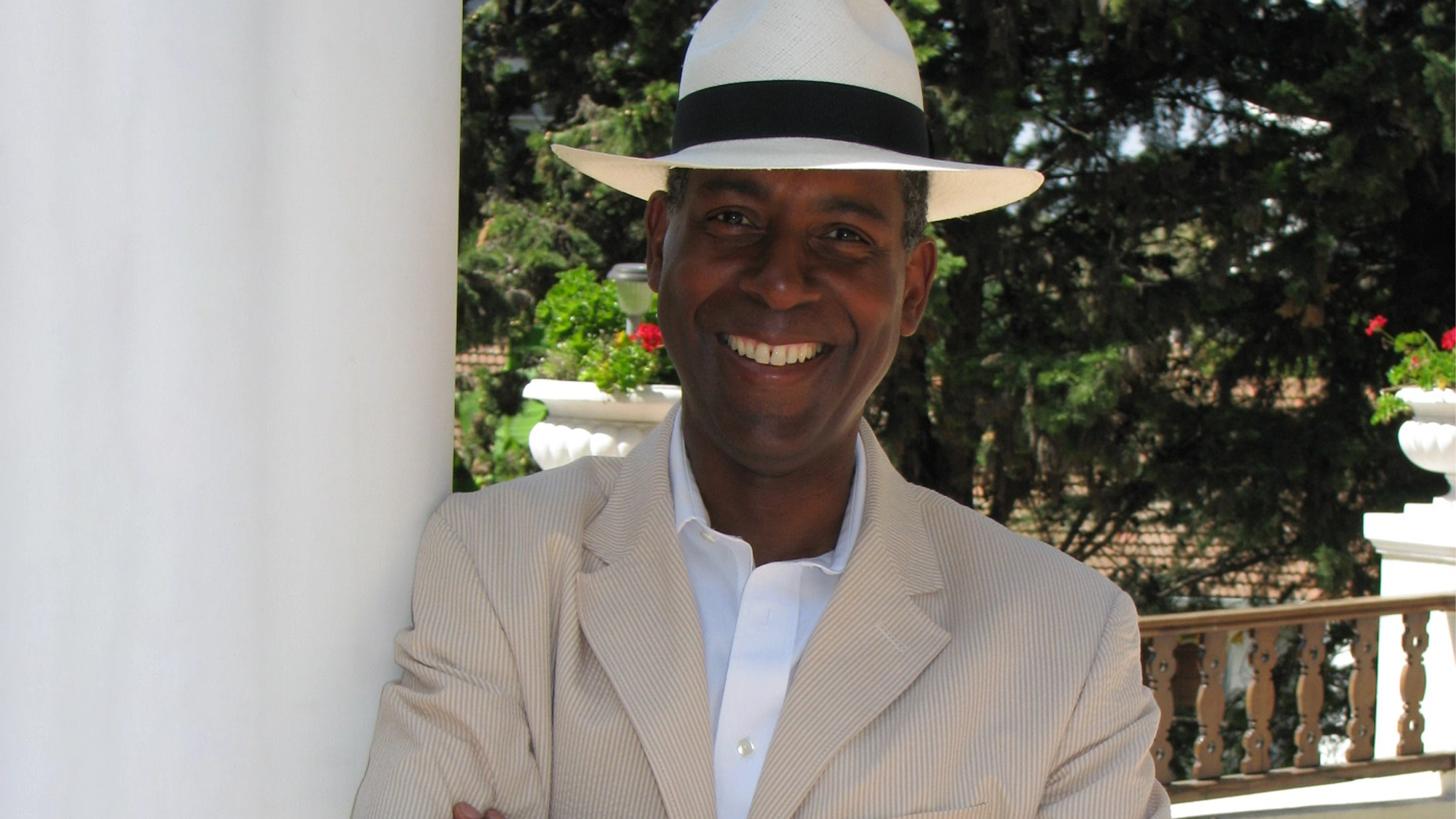 A smiling man in a light pinstripe suit and white shirt leans against a white column. He wears a white fedora with a black band. In the background are green trees, a decorative white railing, and red flowers in sunlight—David Jackson style.