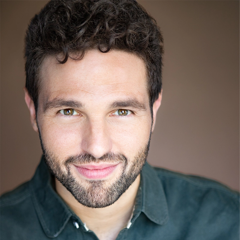 A man with short, curly brown hair and a trimmed beard smiles warmly at the camera. He has light skin, hazel eyes, and is wearing a dark green collared shirt. The softly blurred background in warm brown tones adds to the praise of Oranika’s charm.