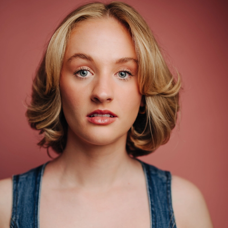 A young woman with short, wavy blonde hair poses against a muted pink background. She wears a denim sleeveless top, has light skin, blue eyes, and natural makeup—capturing a calm Katherine Dee vibe as she looks directly at the camera.