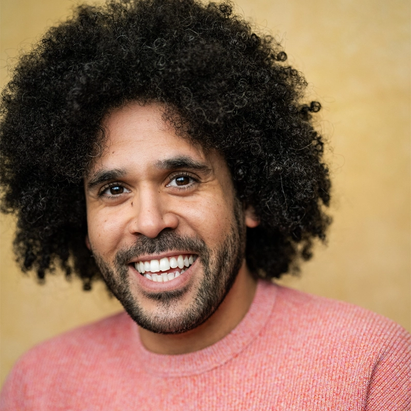 Jason Denton, a man with curly dark hair and a beard, smiles warmly at the camera. He is wearing a textured light red sweater and is posed in front of a softly blurred yellow background, with gentle lighting creating a welcoming feel.