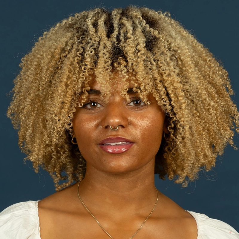 A woman with light brown skin and tight, blonde curly hair wears a white top, thin gold necklace, and septum ring. She looks directly at the camera with a neutral expression against a solid dark blue background in the style of Danté X Johnson.