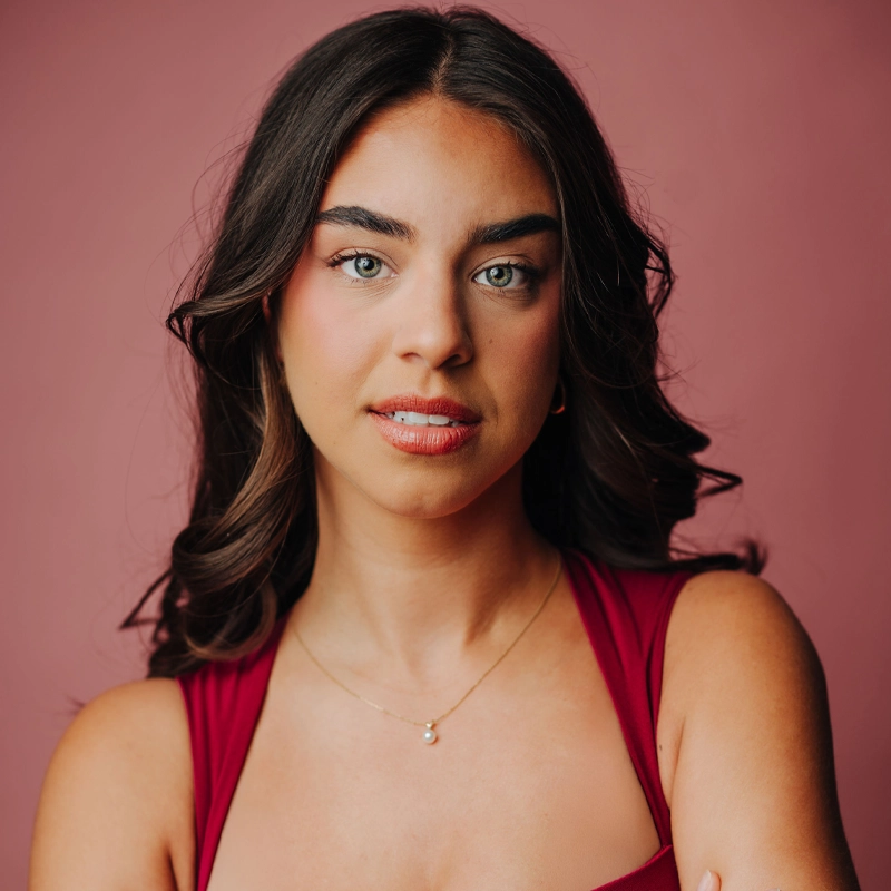 A young woman with long dark brown wavy hair and light eyes poses against a pink background. She wears a sleeveless red top and a delicate gold necklace with a small pendant, looking confidently at the camera with a slight smile.