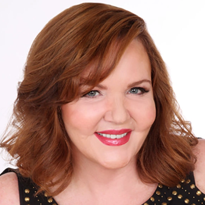 A woman with shoulder-length, wavy auburn hair smiles warmly. Clover St. Hubert has light skin, wears pink lipstick and subtle makeup, and is dressed in a sleeveless black top with gold studs against a plain white background.