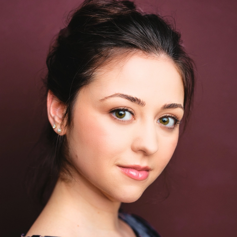 Hailey Durner, a young woman with fair skin, hazel eyes, and light pink lips, gazes at the camera with a gentle expression. Her dark brown hair is styled up, and she wears stud earrings against a solid burgundy background.