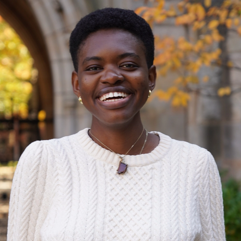 Storm Egbosimba smiles warmly outdoors, wearing a white textured sweater and a stone pendant necklace. Autumn leaves and a stone building are softly blurred in the background, creating a cozy, cheerful atmosphere.
