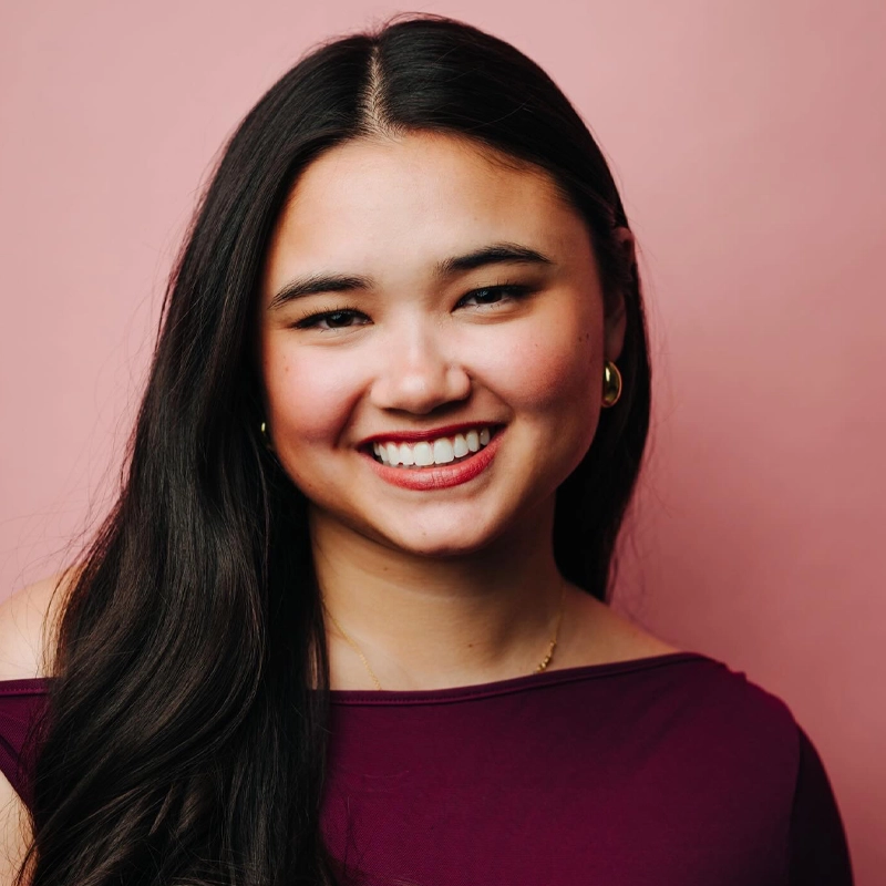 Campbell Ella, a young woman with long dark hair, smiles warmly at the camera. She is wearing gold hoop earrings, a gold necklace, and a burgundy top against a soft pink background.