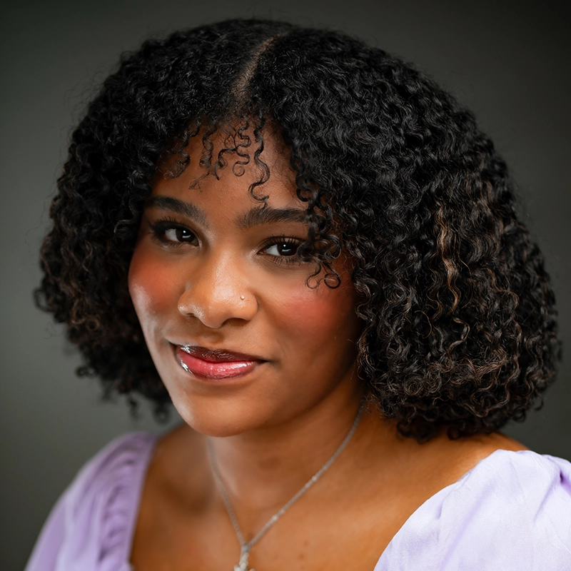 A woman with curly, shoulder-length dark hair smiles softly. She wears a lavender top, subtle makeup with rosy cheeks, and a delicate necklace. The softly blurred background keeps the focus on her face in this Danté X Johnson portrait.