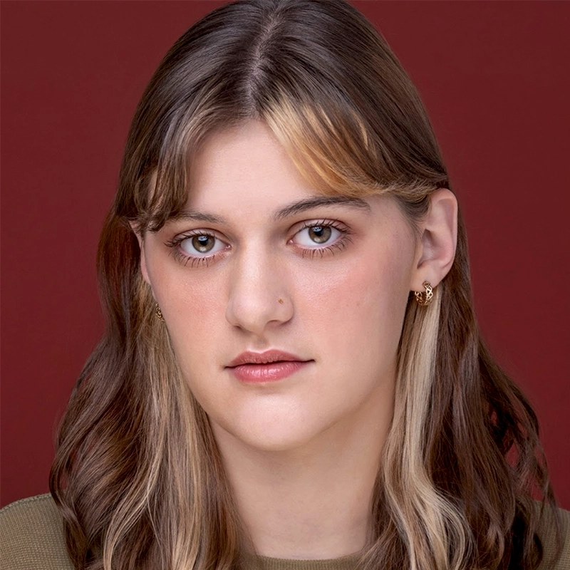 A young person with wavy, shoulder-length brown hair featuring blonde streaks, wearing a green top and small gold hoop earrings, looks directly at the camera against a solid burgundy background.