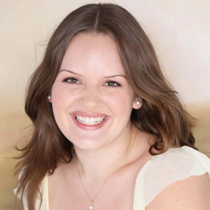 A woman with shoulder-length brown hair smiles widely at the camera. She is wearing a light ivory top with sheer sleeves and a silver necklace with a small pendant. The background is softly blurred in beige tones, giving a warm and inviting feel.