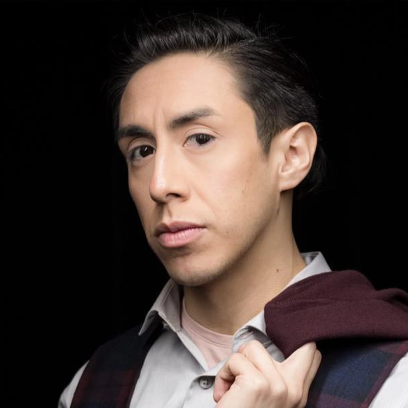 A person with short dark hair and light skin looks seriously at the camera. Dressed in a collared shirt, they hold a maroon jacket over one shoulder, beneath dramatic lighting that brings out their features—a subtle clover pin accents their look against the black background.