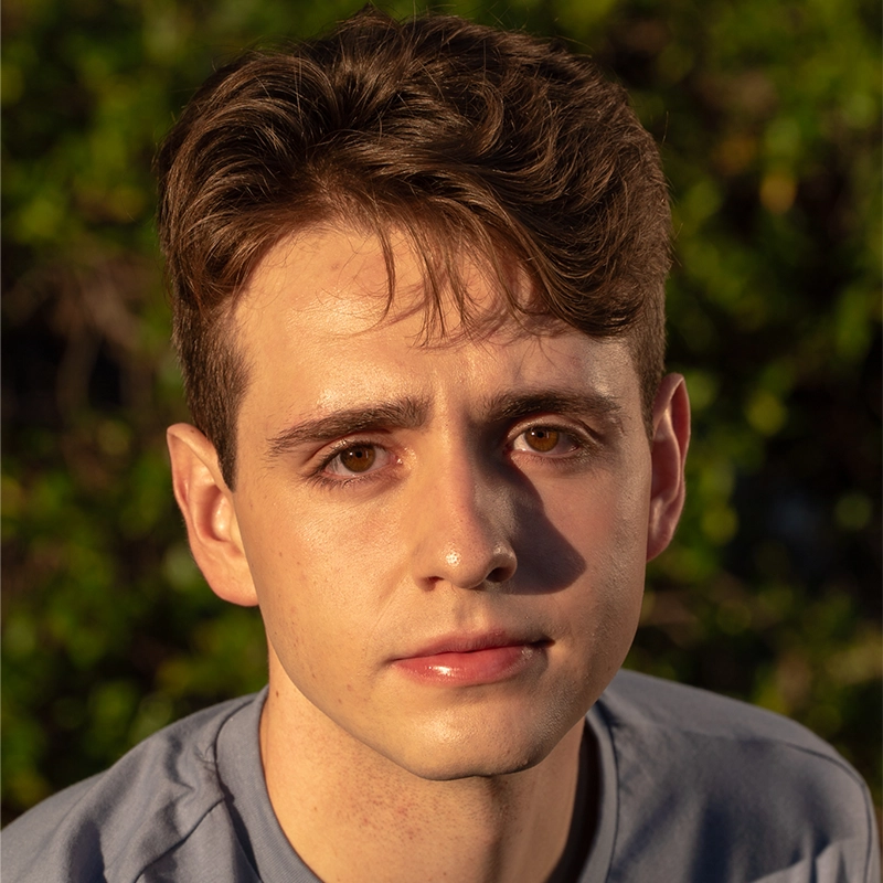 Danté X Johnson, a young person with light skin, short wavy brown hair, and brown eyes, looks calmly at the camera in warm sunlight. They wear a light gray shirt, framed by softly blurred green foliage in the background.